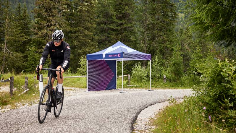 Ein vollflächig bedruckter Faltpavillon von Ecotent Ein Ecotent Faltpavillon, vollflächig bedruckt steht am Waldrand. Ein Fahrradfahrer fährt vorbei.